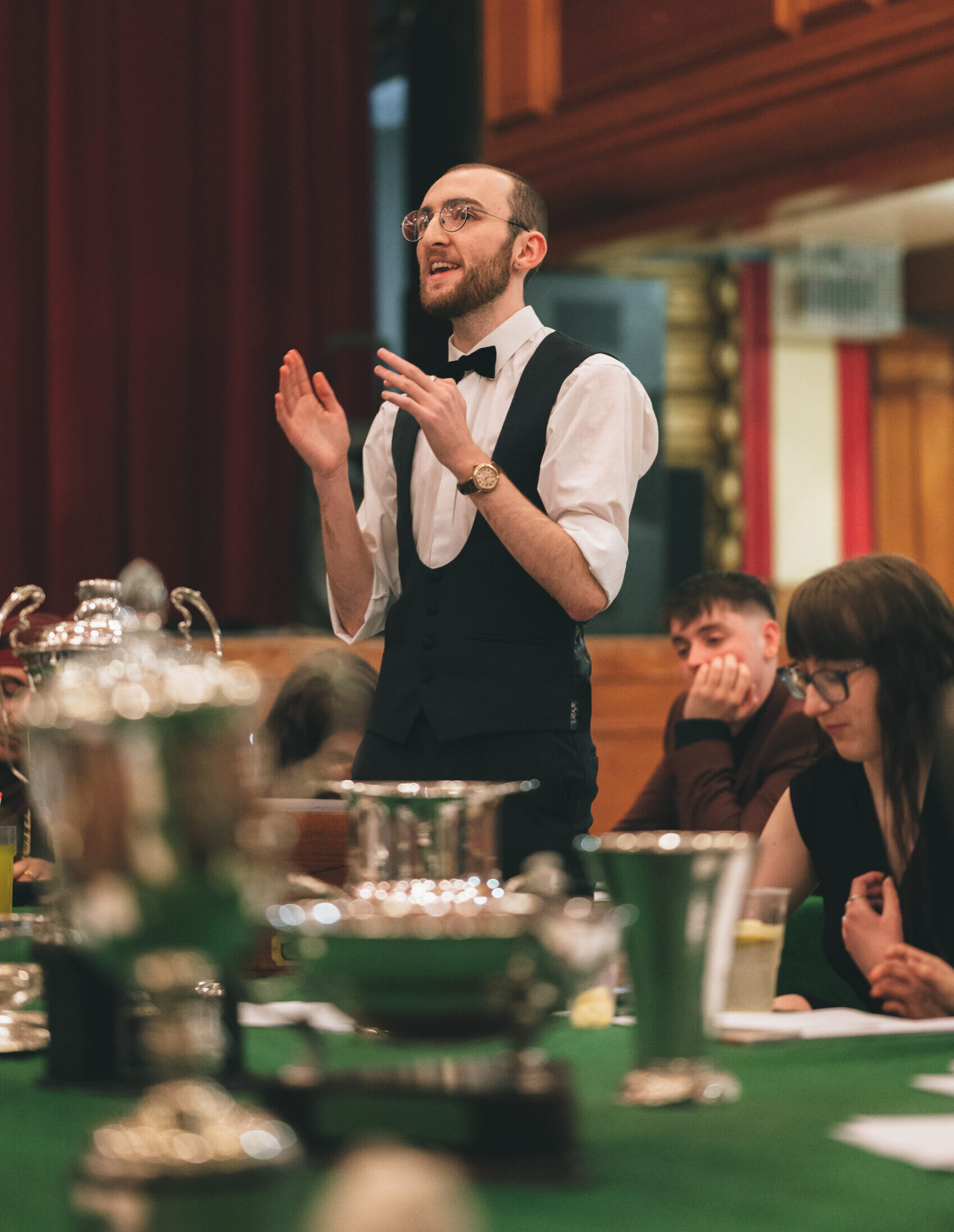 Joseph Hutchison dressed smartly in a dinner suit stands at a table with his arms raised making his point.