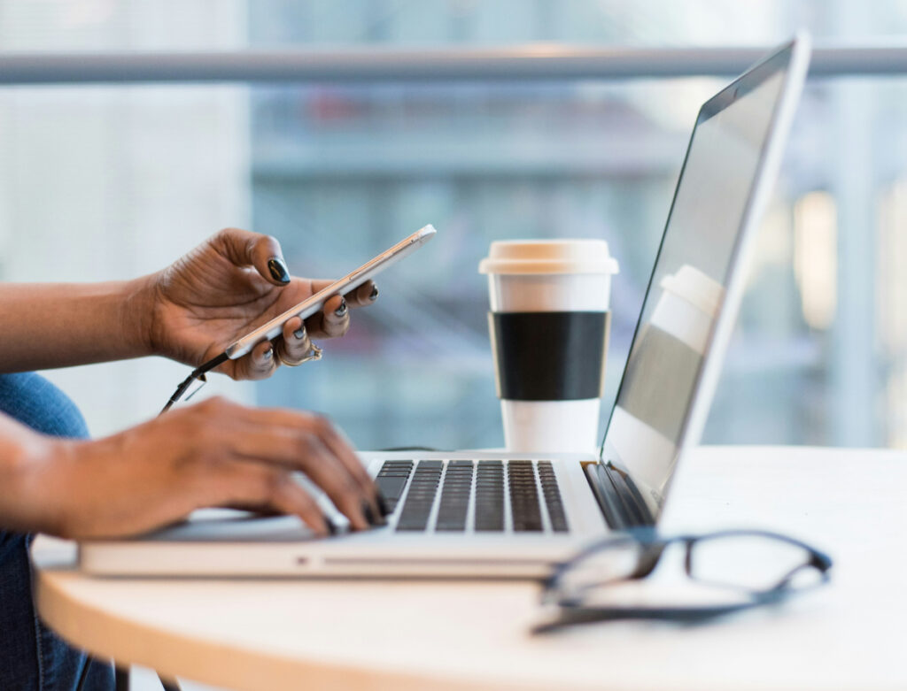 Stock image of a woman sitting at a laptop with her phone in hand.