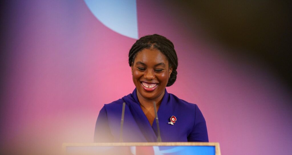 Kemi Badenoch smiling as she stands at a lecture podium.