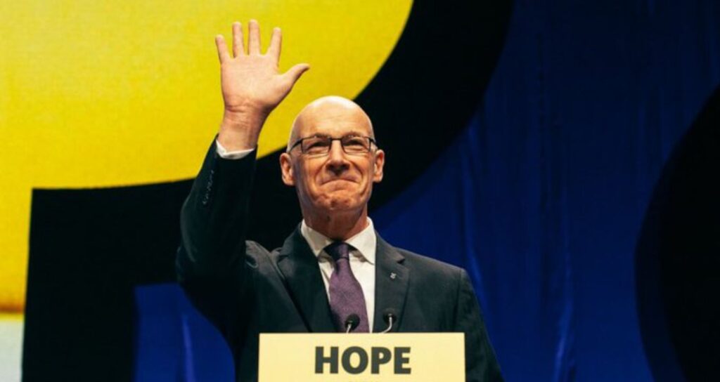 First Minister John Swinney stading at a podium in front of a crowd at the forefront of Scottish politics in 2025.